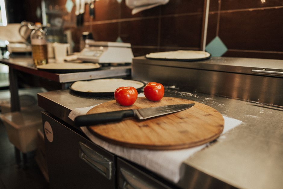 A kitchen scene featuring fresh tomatoes and a knife on a wooden board, set against a counter with pizza dough.