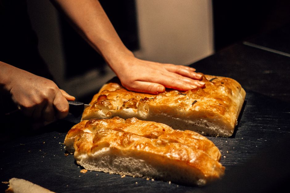 Close-up of hands cutting freshly baked focaccia bread on a dark surface.